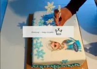 将蓝色雪花贴在蛋糕上。从糖纸上剪下艾尔莎的形象，贴在蛋糕上。蛋糕已经准备就绪，可以等到冰箱里的桌子上...